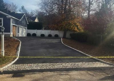 Freshly paved driveway with caution tape across the entrance, bordered by trees and a white fence.