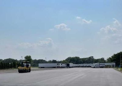 A yellow steamroller is paving a large lot near white trailers and trees under a clear sky.