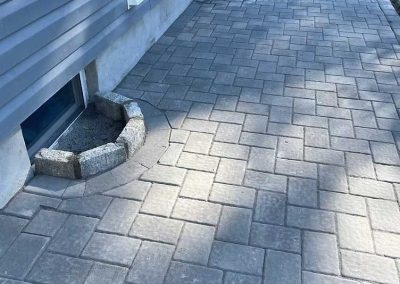 A paved patio with gray bricks beside a house, featuring a small stone barrier around a basement window.