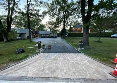 Newly paved driveway with caution tape, trees, and houses in the background at dusk.