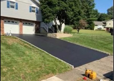 Freshly paved black driveway in front of a suburban house with green lawn and trees.
