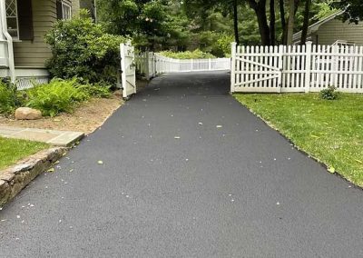 A newly paved driveway leads to a white picket fence beside a house and green lawn.