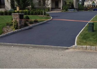Freshly paved driveway with a caution tape barrier, bordered by grass and stone edges, leading to a house.
