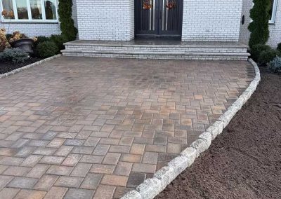 Brick paver driveway bordered with stones leading to double doors of a white brick house.
