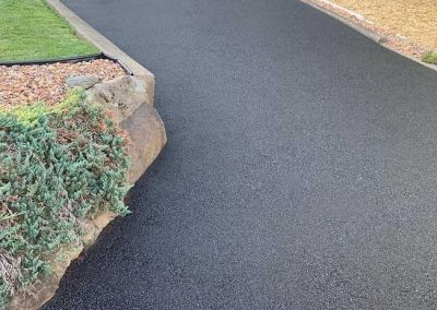 Freshly paved asphalt driveway with caution tape at the bottom, bordered by grass and plants.