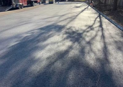 Freshly paved parking lot with tree shadows, trucks, and a few workers in the background under clear sky.