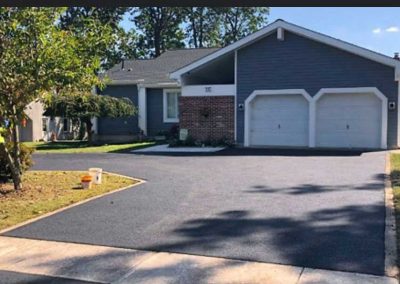 Freshly paved black asphalt driveway in front of a gray house with white trim, two garages, and a tree.