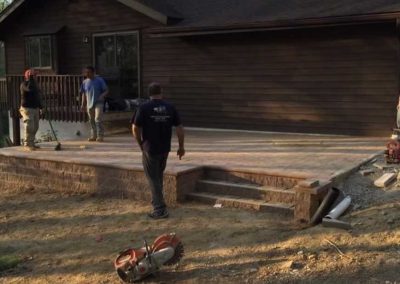 Four workers stand on and around a newly built patio with steps beside a brown wooden house.