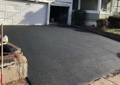 Freshly paved driveway leads to a white garage attached to a green house with a small porch.