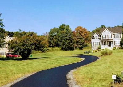 A curving asphalt driveway leads to a white house, surrounded by green grass and some trees.