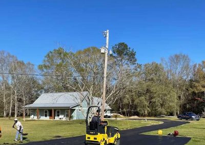 Workers pave a driveway with fresh asphalt near a house; one uses a yellow roller under a clear sky.