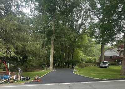 A newly paved driveway leads into a wooded area, with a car parked next to a brick house on the right.