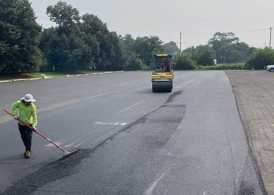 Worker smooths new asphalt with a rake as a steamroller compacts the surface in a large parking lot.