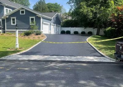 Freshly paved driveway blocked by yellow tape in front of a gray house with a garage, trees, and yard.