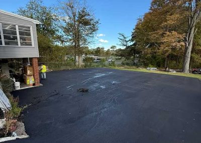 Freshly paved black asphalt driveway next to a house, with trees and blue sky in the background.