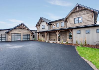 Modern rustic two-story house with attached garage, wooden siding, and large windows on a paved driveway.