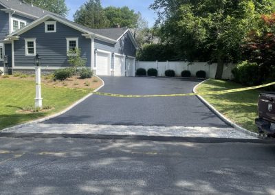 Freshly paved driveway with caution tape in front of a gray house, trees and shrubs in the background.