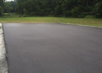 Newly paved asphalt surface with trees and grass in the background under a cloudy sky.
