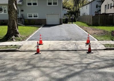 Four orange cones block the entrance to a driveway with fresh asphalt in front of a gray house.