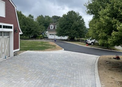 A newly paved stone driveway leads to a road in a suburban neighborhood with green trees and cloudy skies.