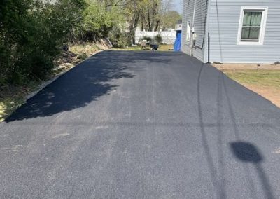 Freshly paved asphalt driveway beside a gray house with trees and a blue sky in the background.